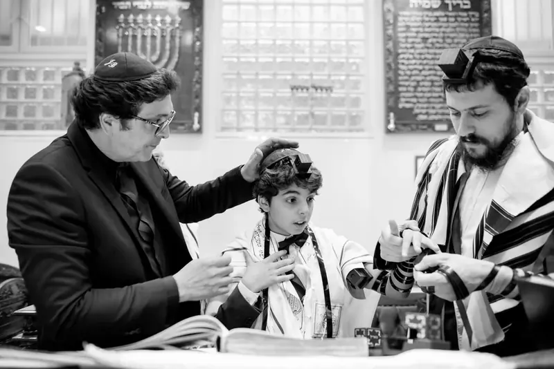 Photo en noir et blanc d'un jeune Bar Mitzvah qui met ses téfilines, un adulte la main posée sur sa tête et un autre l'aidant à enrouler la lanière